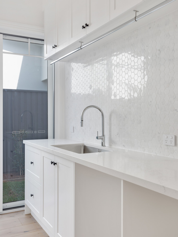 Inspiration for a large beach style galley dedicated laundry room in Brisbane with an undermount sink, shaker cabinets, white cabinets, marble benchtops, white splashback, marble splashback, white walls, light hardwood floors, a side-by-side washer and dryer, brown floor and white benchtop.