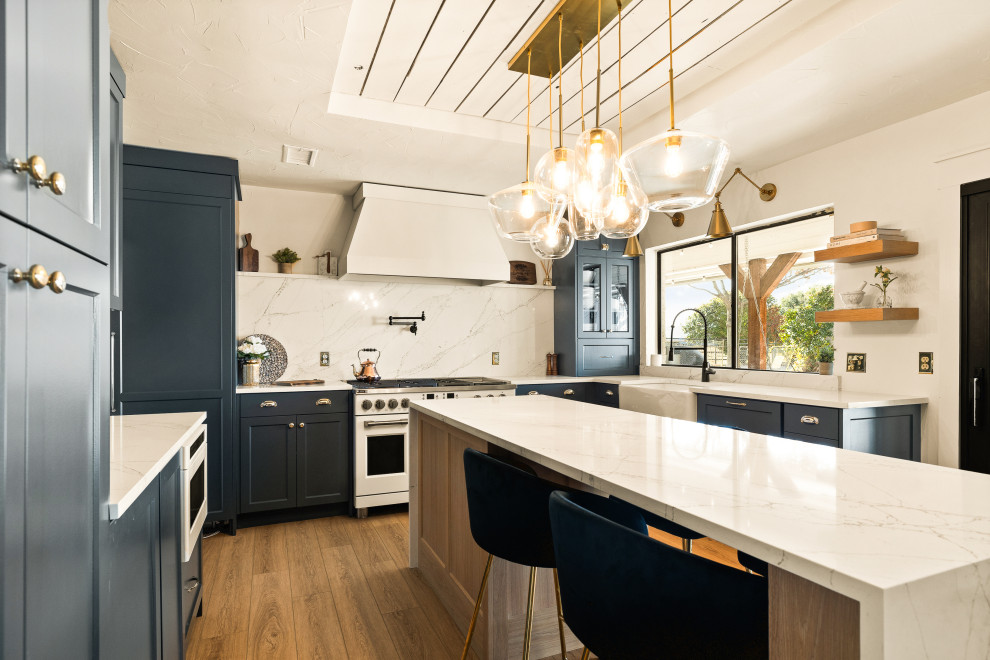 Photo of a transitional u-shaped kitchen in Dallas with a farmhouse sink, shaker cabinets, blue cabinets, white splashback, stone slab splashback, white appliances, medium hardwood floors, with island, brown floor, white benchtop and recessed.