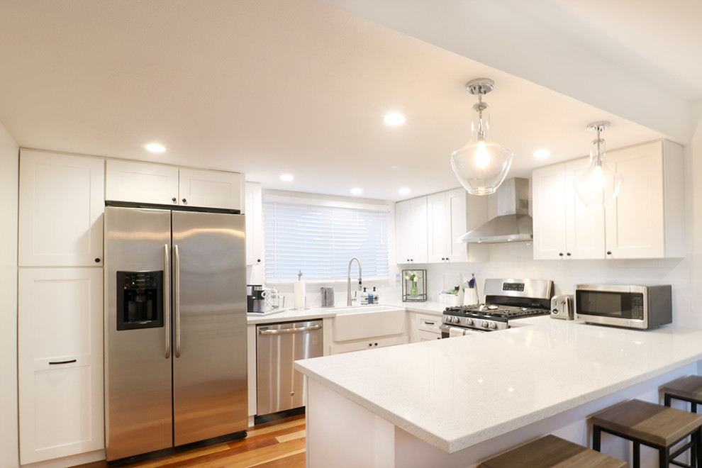This is an example of a small transitional u-shaped open plan kitchen in Orange County with a drop-in sink, recessed-panel cabinets, white cabinets, marble benchtops, white splashback, ceramic splashback, stainless steel appliances, medium hardwood floors, no island and brown floor.