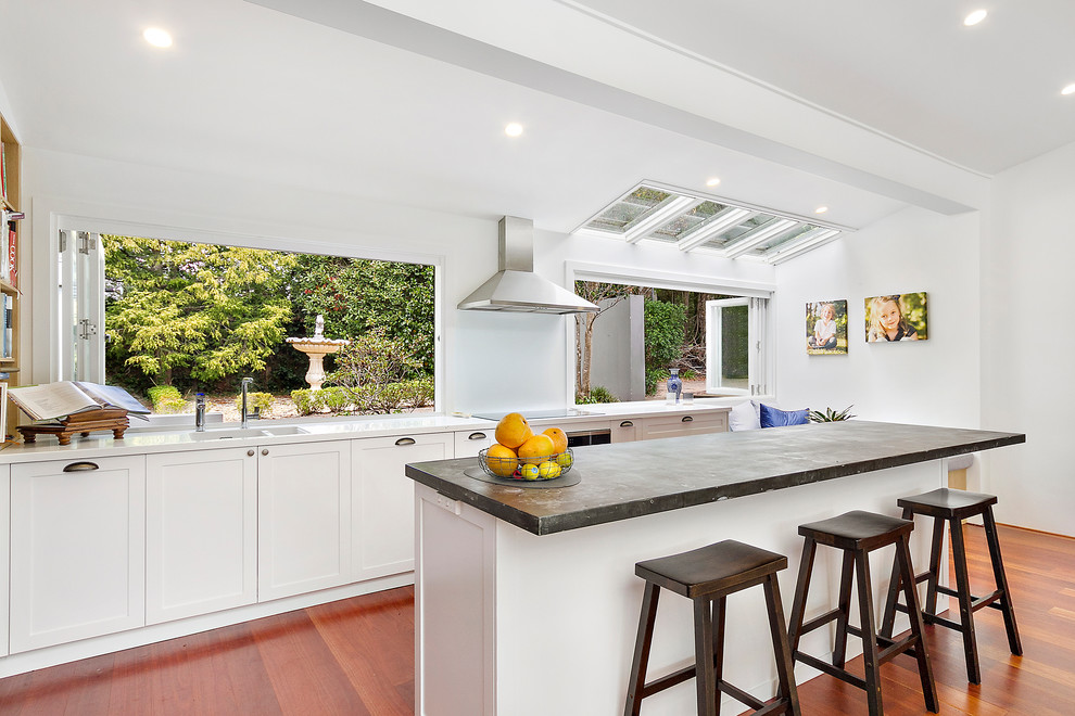 This is an example of an expansive contemporary l-shaped open plan kitchen in Sydney with an integrated sink, shaker cabinets, white cabinets, zinc benchtops, white splashback, glass sheet splashback, black appliances, light hardwood floors, with island, brown floor and grey benchtop.
