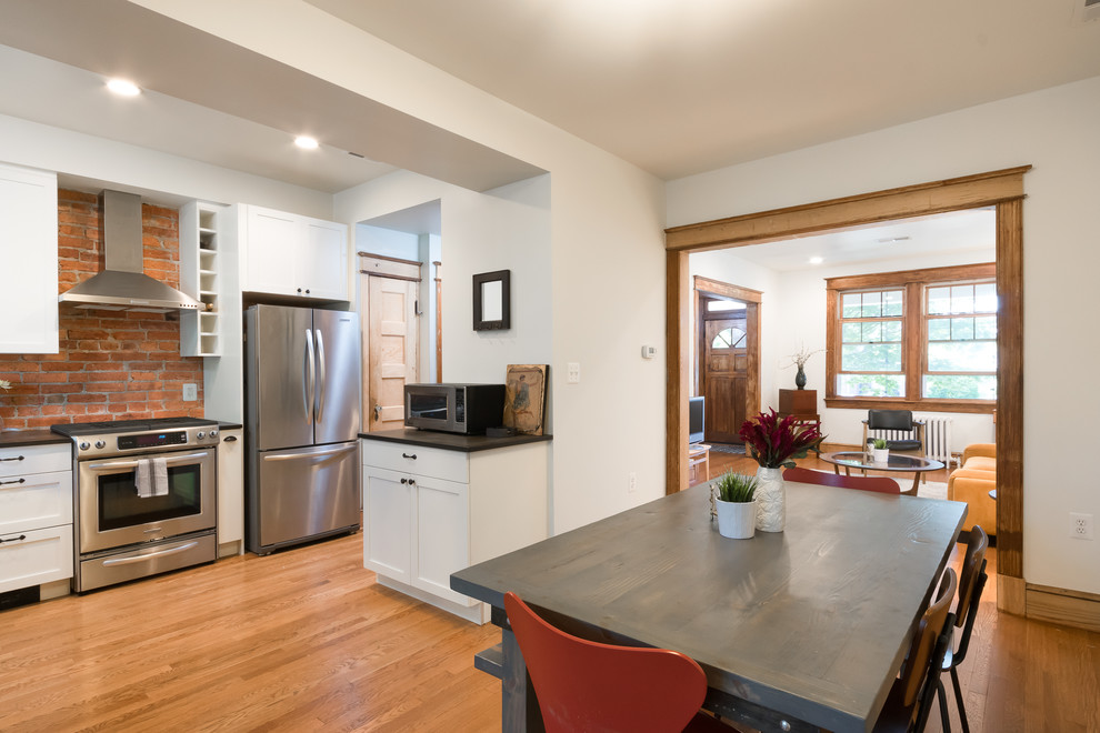 Mid-sized arts and crafts l-shaped eat-in kitchen in DC Metro with shaker cabinets, white cabinets, medium hardwood floors, no island, brown floor, black benchtop, an undermount sink, red splashback, brick splashback and stainless steel appliances.