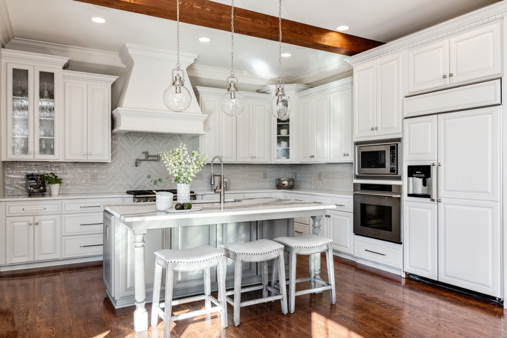 Photo of a large traditional l-shaped kitchen in Charlotte with an undermount sink, raised-panel cabinets, white cabinets, quartz benchtops, grey splashback, porcelain splashback, stainless steel appliances, dark hardwood floors, with island, brown floor and white benchtop.