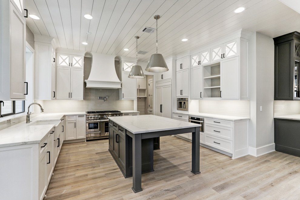 This is an example of a large transitional u-shaped open plan kitchen in Houston with a farmhouse sink, recessed-panel cabinets, white cabinets, granite benchtops, grey splashback, glass tile splashback, stainless steel appliances, light hardwood floors, with island, multi-coloured floor, white benchtop and timber.