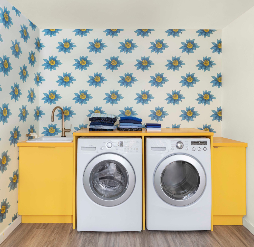 ポートランドにあるお手頃価格の中くらいなミッドセンチュリースタイルのおしゃれな家事室 (シングルシンク、フラットパネル扉のキャビネット、黄色いキャビネット、ラミネートカウンター、青い壁、ラミネートの床、左右配置の洗濯機・乾燥機、茶色い床、黄色いキッチンカウンター、壁紙) の写真