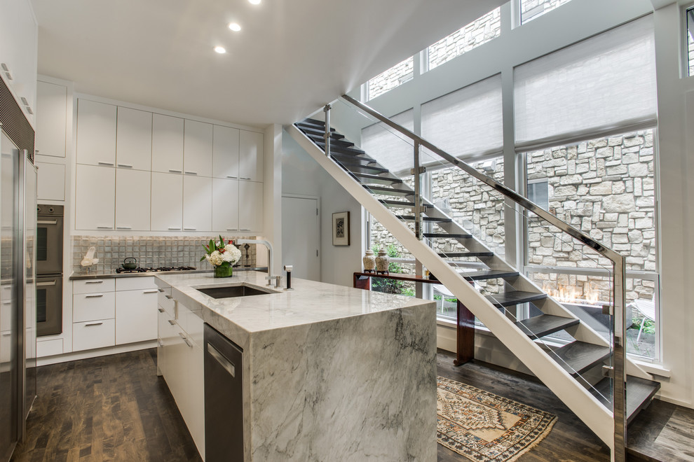 Example of a mid-sized minimalist l-shaped medium tone wood floor open concept kitchen design in Dallas with an undermount sink, flat-panel cabinets, white cabinets, quartzite countertops, glass tile backsplash, stainless steel appliances, an island and gray backsplash