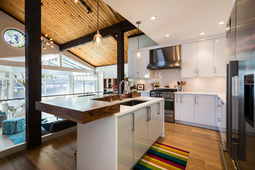 Example of a mid-sized l-shaped light wood floor and brown floor open concept kitchen design in Vancouver with an undermount sink, flat-panel cabinets, white cabinets, quartz countertops, white backsplash, ceramic backsplash, stainless steel appliances and an island