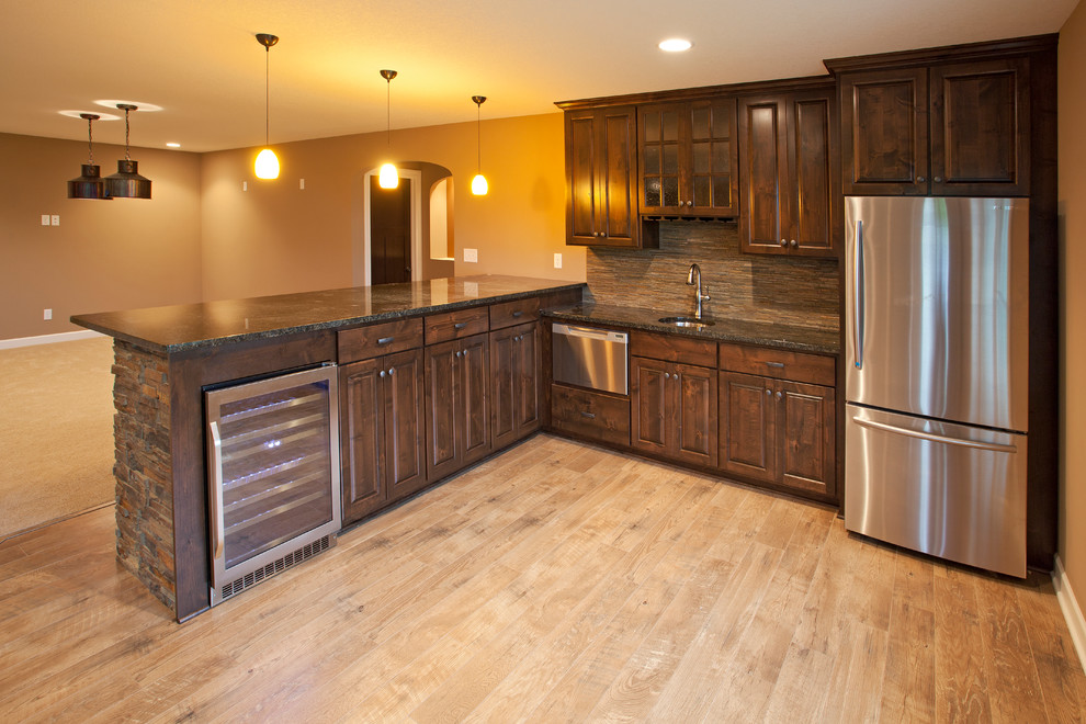 Photo of a mid-sized traditional l-shaped wet bar in Minneapolis with an undermount sink, raised-panel cabinets, medium wood cabinets, granite benchtops, brown splashback, stone tile splashback, light hardwood floors, brown floor and black benchtop.
