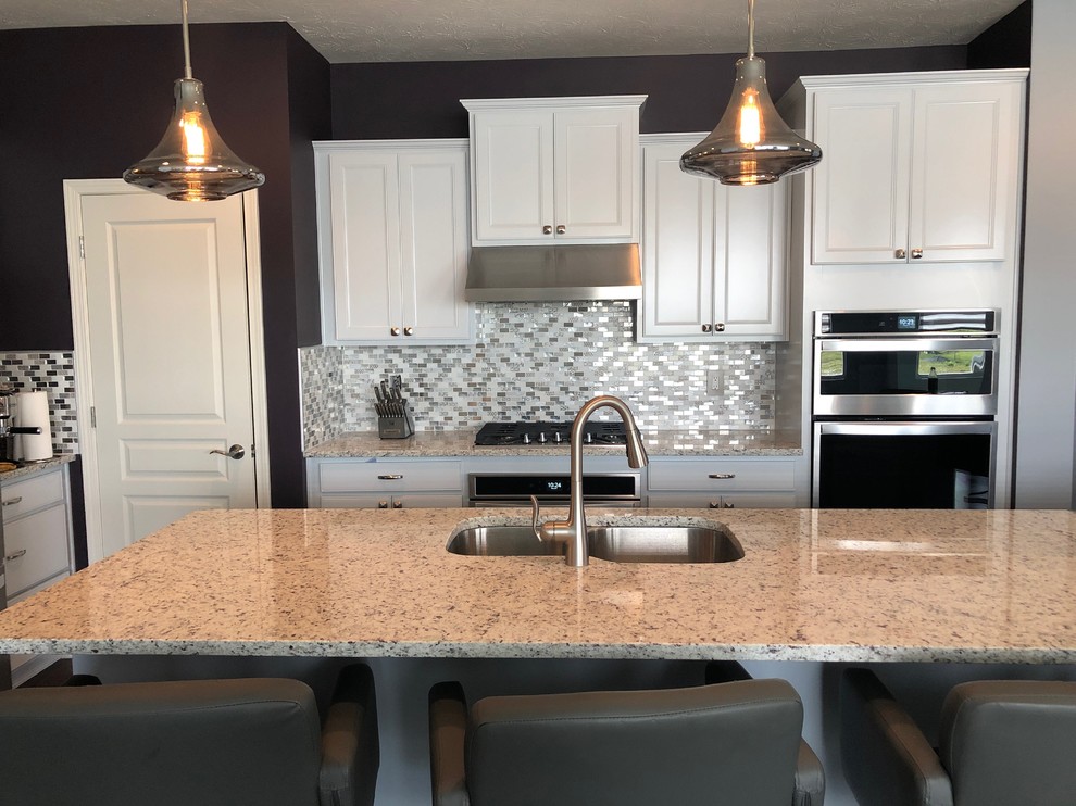 Small traditional l-shaped open plan kitchen in Indianapolis with recessed-panel cabinets, white cabinets, granite benchtops, multi-coloured splashback, mosaic tile splashback, stainless steel appliances, vinyl floors, with island, brown floor and white benchtop.