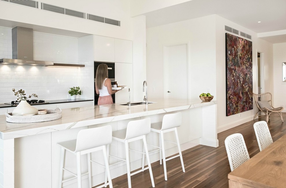 Photo of a mid-sized beach style galley eat-in kitchen in Sunshine Coast with a double-bowl sink, white cabinets, quartz benchtops, white splashback, subway tile splashback, stainless steel appliances, medium hardwood floors, with island and black benchtop.