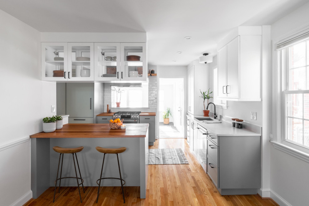 This is an example of a mid-sized transitional l-shaped eat-in kitchen in DC Metro with an undermount sink, shaker cabinets, green cabinets, wood benchtops, white splashback, subway tile splashback, stainless steel appliances, light hardwood floors, a peninsula, brown floor and brown benchtop.