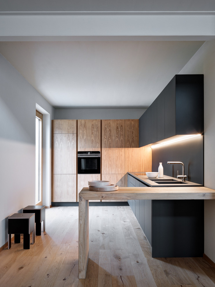 Design ideas for a modern l-shaped separate kitchen in Milan with flat-panel cabinets, black cabinets, wood benchtops, black splashback, black appliances, medium hardwood floors, brown floor, an integrated sink, no island and grey benchtop.