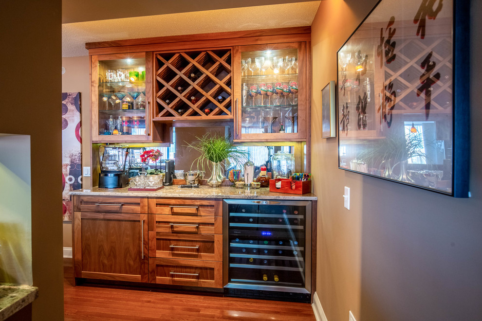 Small traditional single-wall home bar in Chicago with recessed-panel cabinets, medium wood cabinets, granite benchtops, mirror splashback, dark hardwood floors, brown floor and multi-coloured benchtop.