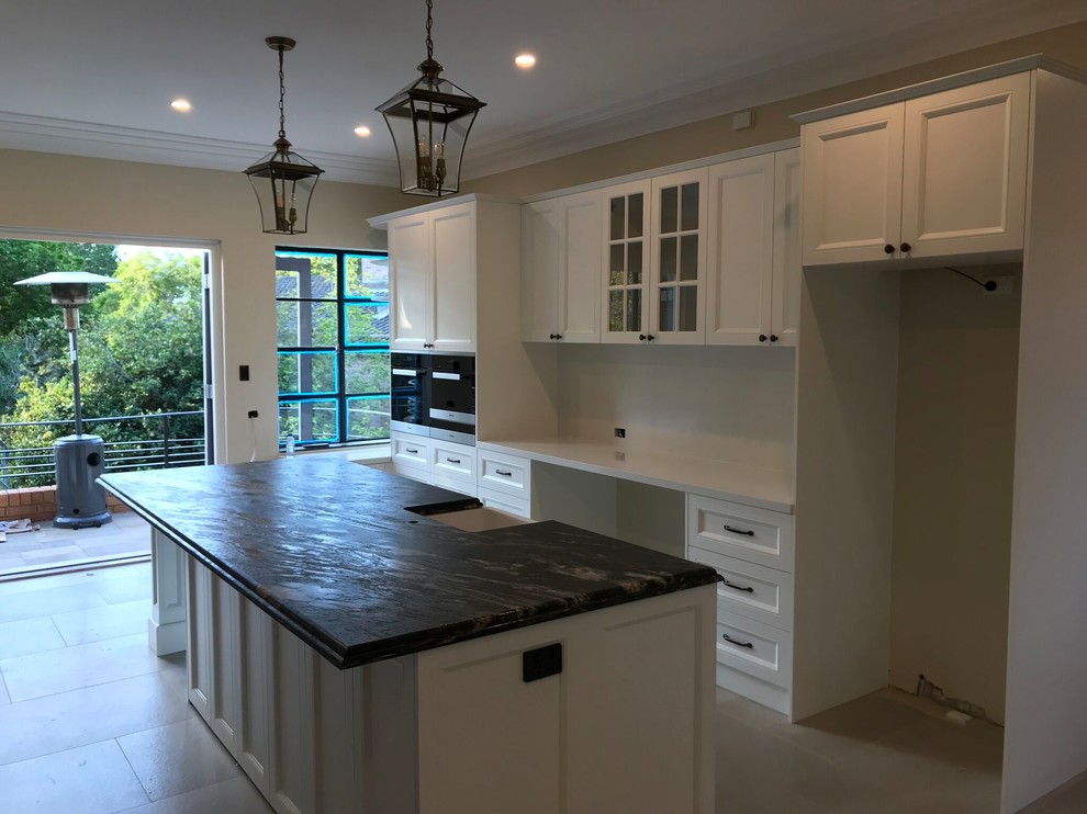 Inspiration for an expansive traditional galley open plan kitchen in Sydney with an undermount sink, shaker cabinets, white cabinets, granite benchtops, white splashback, stone slab splashback, black appliances, multiple islands and black benchtop.