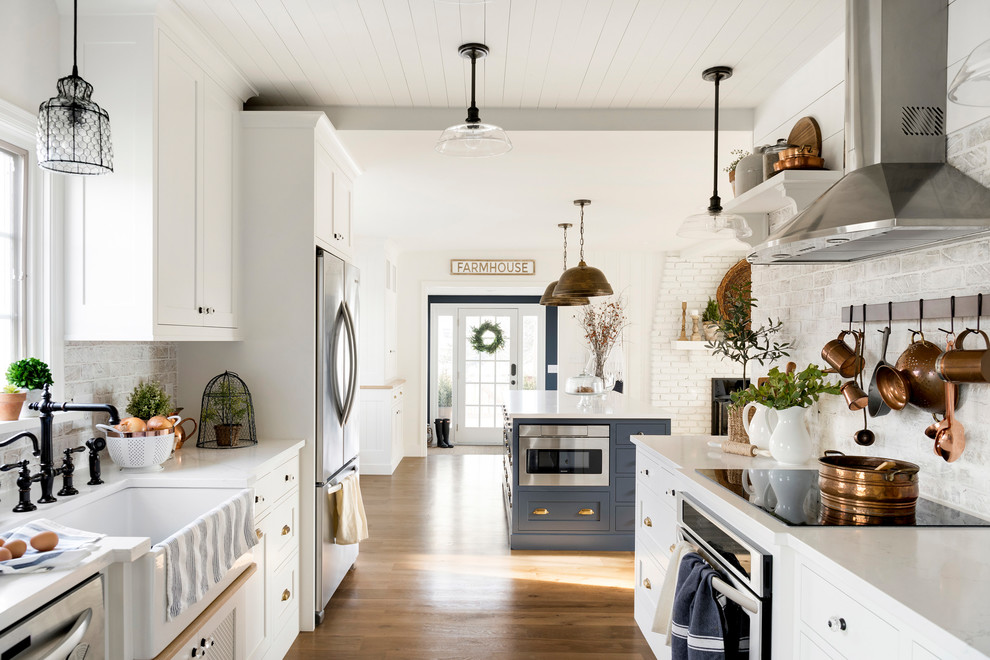 Country kitchen in Minneapolis with a farmhouse sink, white cabinets, brick splashback, stainless steel appliances, with island, white benchtop, shaker cabinets, grey splashback and medium hardwood floors.