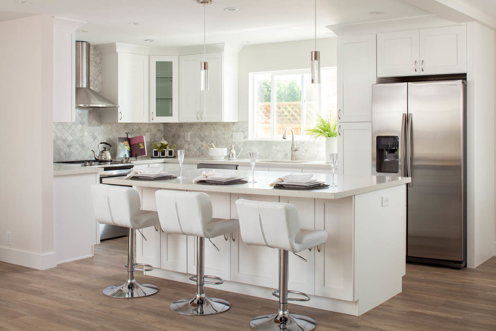 Inspiration for a mid-sized arts and crafts u-shaped open plan kitchen in San Francisco with an undermount sink, shaker cabinets, white cabinets, quartz benchtops, grey splashback, marble splashback, stainless steel appliances, laminate floors, with island, brown floor and white benchtop.