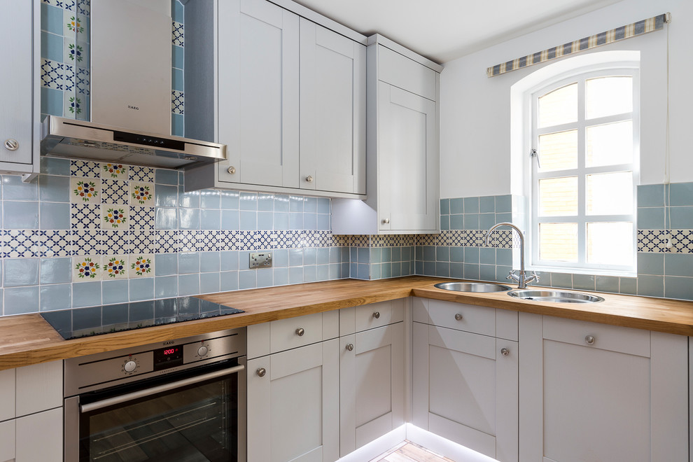 This is an example of a mid-sized traditional u-shaped eat-in kitchen in London with a double-bowl sink, raised-panel cabinets, grey cabinets, wood benchtops, blue splashback, mosaic tile splashback, black appliances, cement tiles and no island.