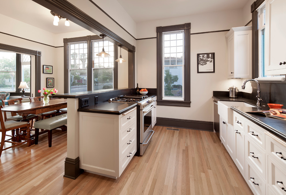 Design ideas for a small traditional galley eat-in kitchen in Santa Barbara with a farmhouse sink, recessed-panel cabinets, white cabinets, granite benchtops, black splashback, stainless steel appliances and light hardwood floors.