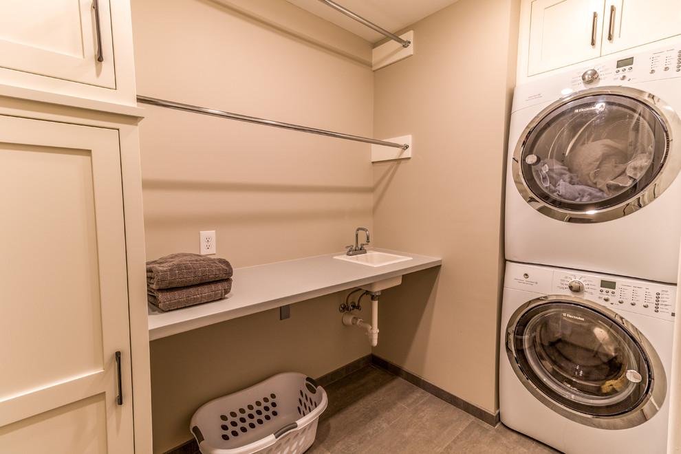 This is an example of a large transitional l-shaped dedicated laundry room in Minneapolis with an utility sink, recessed-panel cabinets, white cabinets, marble benchtops, beige walls, porcelain floors, a stacked washer and dryer, grey floor and grey benchtop.
