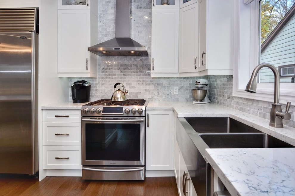 Example of a mid-sized transitional l-shaped medium tone wood floor and brown floor eat-in kitchen design in Toronto with a farmhouse sink, shaker cabinets, white cabinets, quartz countertops, gray backsplash, marble backsplash, stainless steel appliances, an island and white countertops