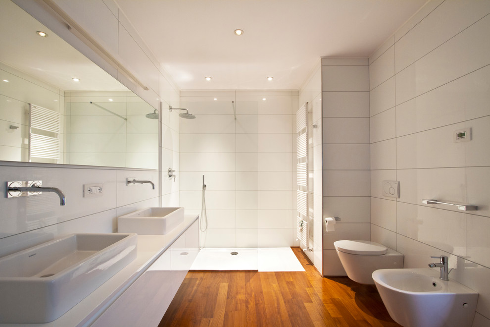 Inspiration for a large contemporary 3/4 bathroom in Other with flat-panel cabinets, white cabinets, a curbless shower, white tile, medium hardwood floors, a wall-mount toilet and a vessel sink.