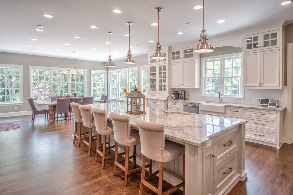 Inspiration for a large transitional l-shaped medium tone wood floor eat-in kitchen remodel with a farmhouse sink, shaker cabinets, white cabinets, white backsplash, subway tile backsplash, stainless steel appliances, an island and marble countertops