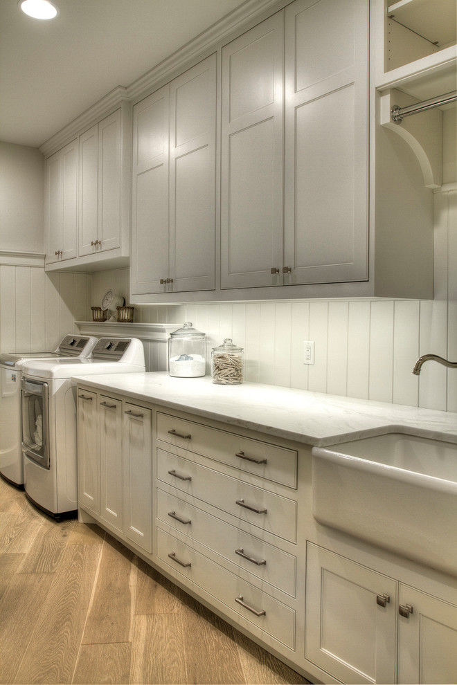 Design ideas for a large traditional galley dedicated laundry room in Minneapolis with a farmhouse sink, recessed-panel cabinets, white cabinets, marble benchtops, white walls, light hardwood floors, a side-by-side washer and dryer and white benchtop.