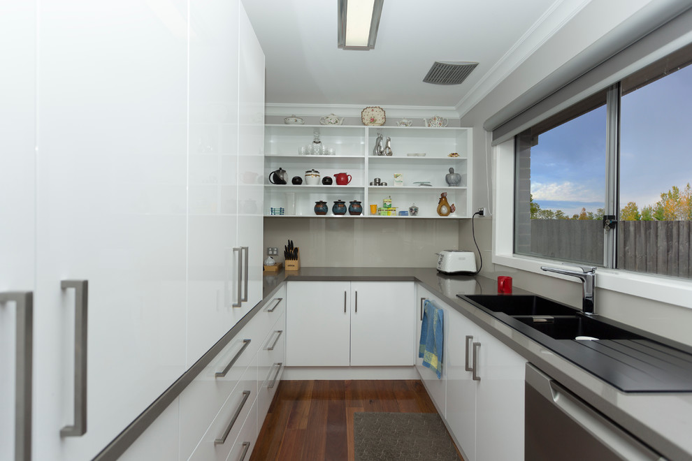 Photo of a small contemporary u-shaped kitchen pantry in Melbourne with a double-bowl sink, flat-panel cabinets, white cabinets, quartz benchtops, beige splashback, glass sheet splashback, stainless steel appliances, medium hardwood floors, no island, brown floor and grey benchtop.