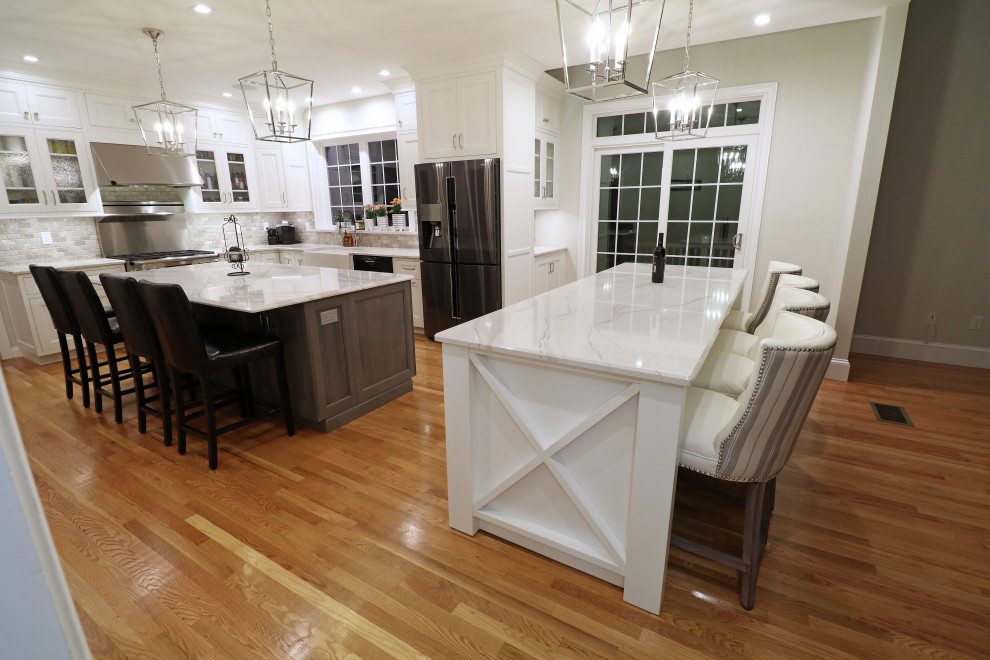 Inspiration for a large transitional l-shaped kitchen in Boston with a farmhouse sink, beaded inset cabinets, white cabinets, quartz benchtops, grey splashback, travertine splashback, stainless steel appliances, medium hardwood floors, multiple islands, yellow floor and white benchtop.