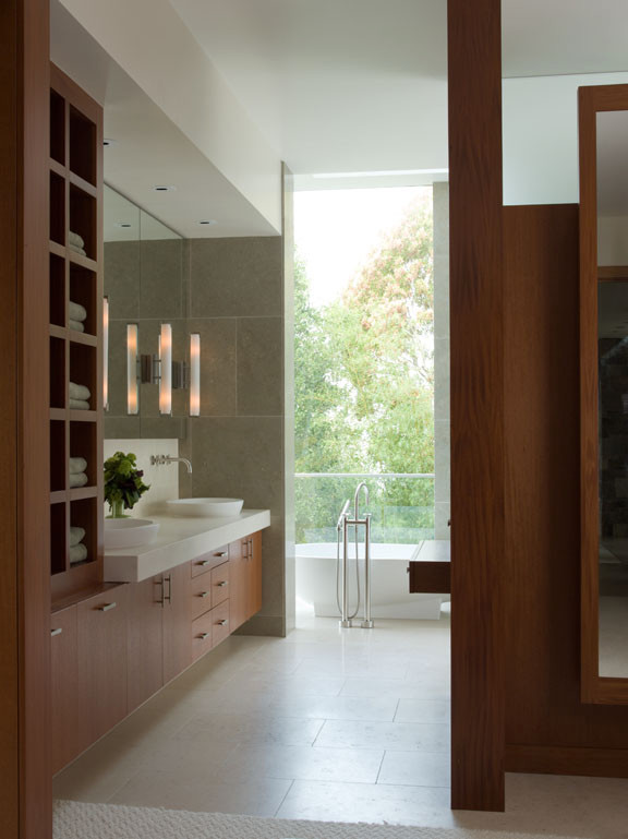 На фото: большая главная ванная комната в стиле модернизм с плоскими фасадами, фасадами цвета дерева среднего тона, отдельно стоящей ванной, душем без бортиков, унитазом-моноблоком, плиткой из известняка, белыми стенами, полом из известняка, настольной раковиной, столешницей из известняка, бежевым полом, душем с распашными дверями, белой столешницей, тумбой под две раковины и встроенной тумбой с