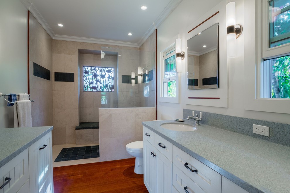 This is an example of a mid-sized beach style 3/4 bathroom in Hawaii with furniture-like cabinets, white cabinets, a curbless shower, a two-piece toilet, beige tile, porcelain tile, white walls, dark hardwood floors, an undermount sink, engineered quartz benchtops, brown floor and an open shower.