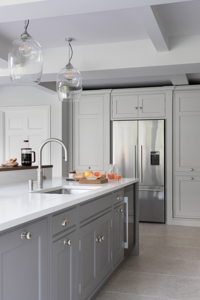 Large classic grey and white l-shaped open plan kitchen in Surrey with a built-in sink, shaker cabinets, grey cabinets, quartz worktops, metallic splashback, mirror splashback, stainless steel appliances, limestone flooring, an island, grey floors, white worktops and exposed beams.
