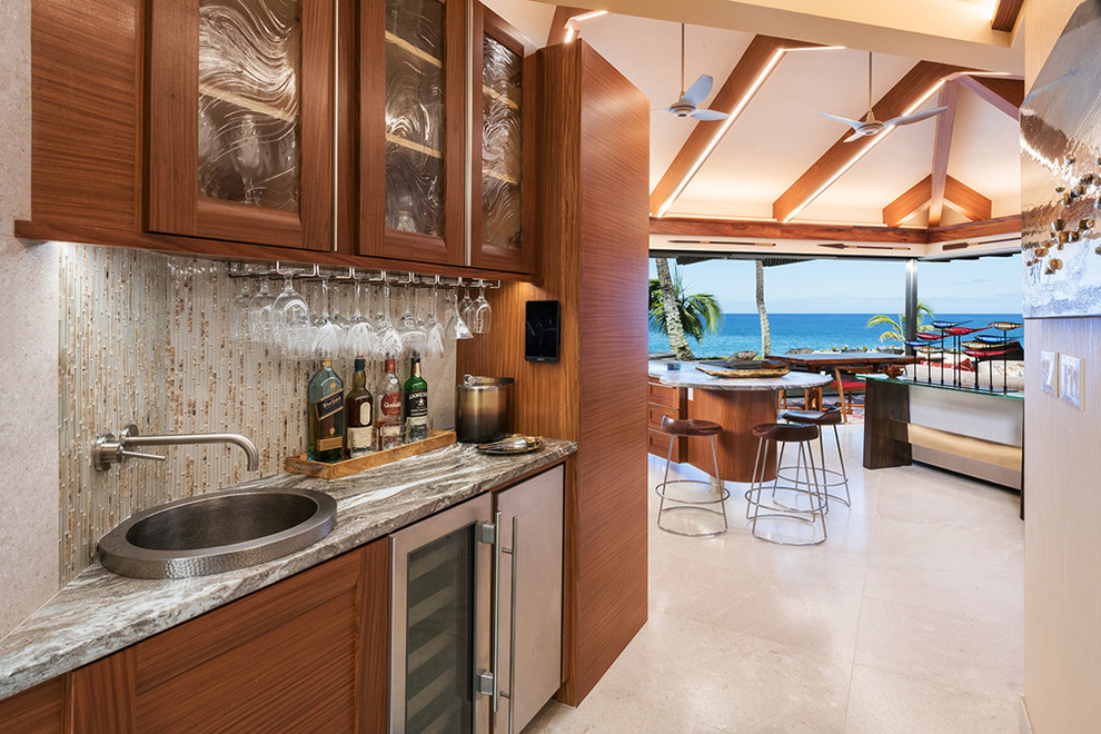 Tropical single-wall wet bar in Hawaii with a drop-in sink, glass-front cabinets, medium wood cabinets, granite benchtops, beige splashback, glass tile splashback, limestone floors, beige floor and brown benchtop.
