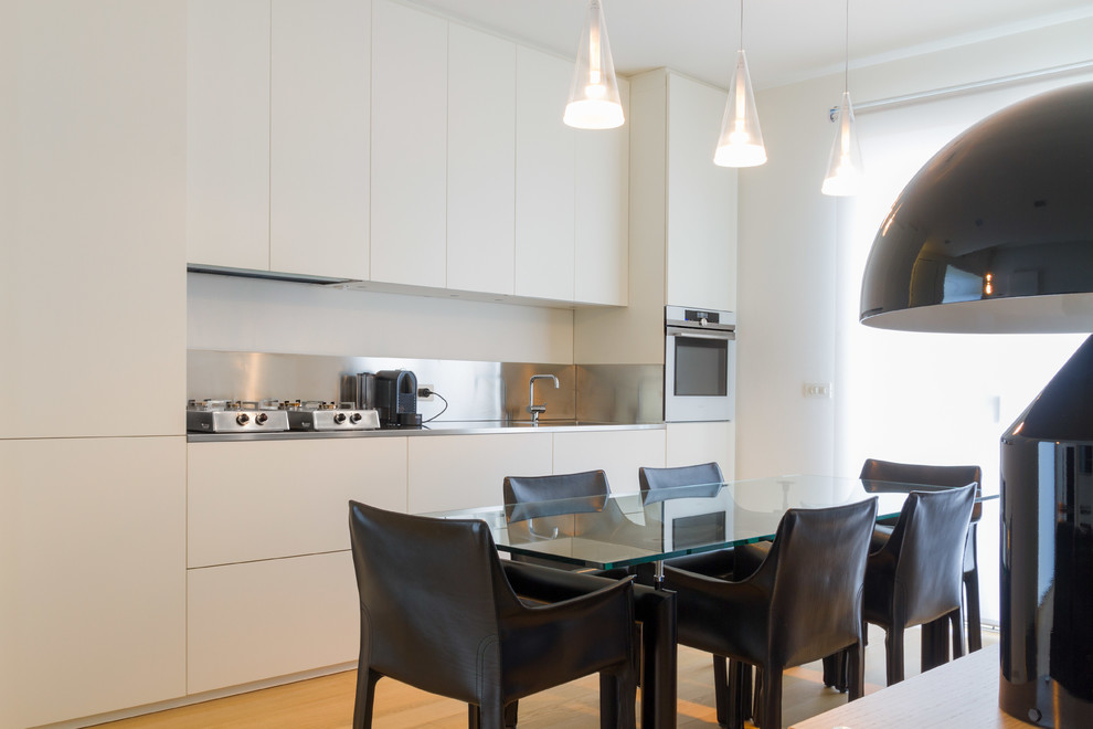 This is an example of a contemporary single-wall kitchen in Bologna with flat-panel cabinets, white cabinets, stainless steel appliances, an undermount sink, stainless steel benchtops, grey splashback, medium hardwood floors, brown floor and grey benchtop.