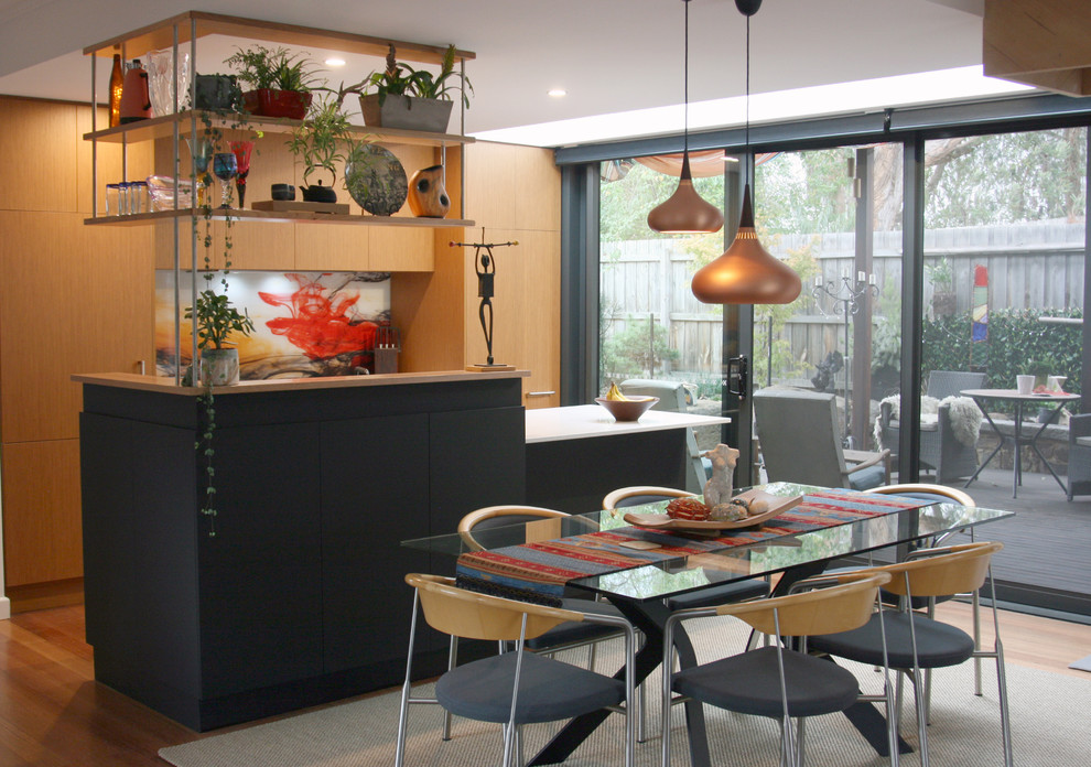 This is an example of a small midcentury galley open plan kitchen in Melbourne with a double-bowl sink, flat-panel cabinets, medium wood cabinets, solid surface benchtops, multi-coloured splashback, glass sheet splashback, stainless steel appliances, medium hardwood floors, with island, brown floor and white benchtop.