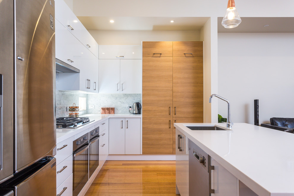 This is an example of a mid-sized contemporary u-shaped open plan kitchen in Melbourne with an undermount sink, solid surface benchtops, white splashback, stone tile splashback, stainless steel appliances, medium hardwood floors, with island, white benchtop, flat-panel cabinets, white cabinets and brown floor.