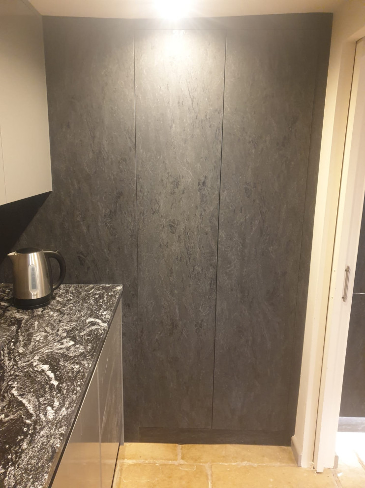 Photo of a mid-sized contemporary l-shaped laundry room in West Midlands with flat-panel cabinets, granite benchtops, black splashback, engineered quartz splashback, black benchtop, a drop-in sink, black cabinets, travertine floors, beige floor and vaulted.