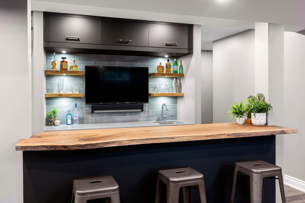 This is an example of a large country galley wet bar in Ottawa with an undermount sink, wood benchtops, flat-panel cabinets, black cabinets, grey splashback, ceramic splashback, laminate floors, multi-coloured floor and grey benchtop.
