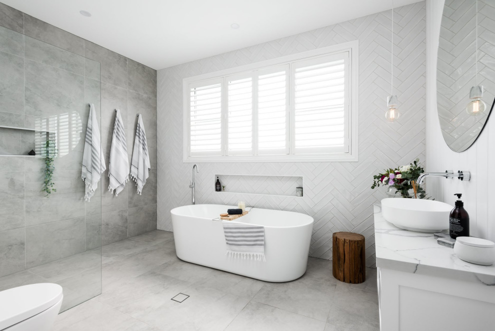 Пример оригинального дизайна: ванная комната в стиле кантри с белыми фасадами, отдельно стоящей ванной, душем без бортиков, белой плиткой, настольной раковиной, серым полом, открытым душем, белой столешницей, нишей и тумбой под одну раковину