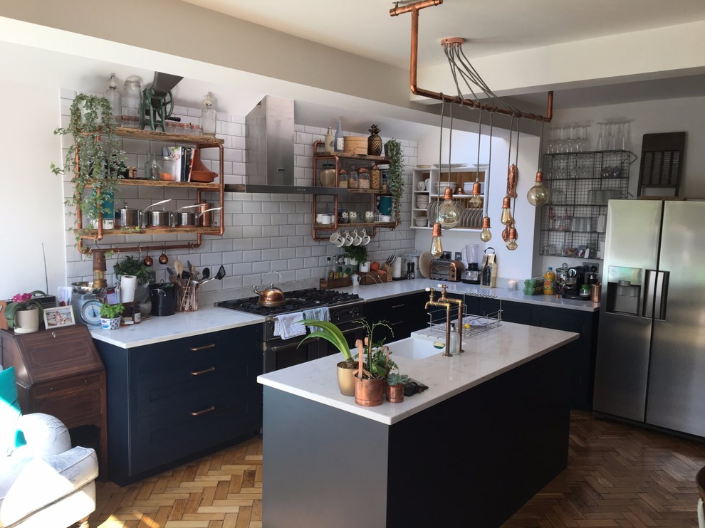 Photo of a small industrial l-shaped open plan kitchen in London with a farmhouse sink, shaker cabinets, blue cabinets, quartzite benchtops, white splashback, porcelain splashback, stainless steel appliances, medium hardwood floors, with island, brown floor and white benchtop.