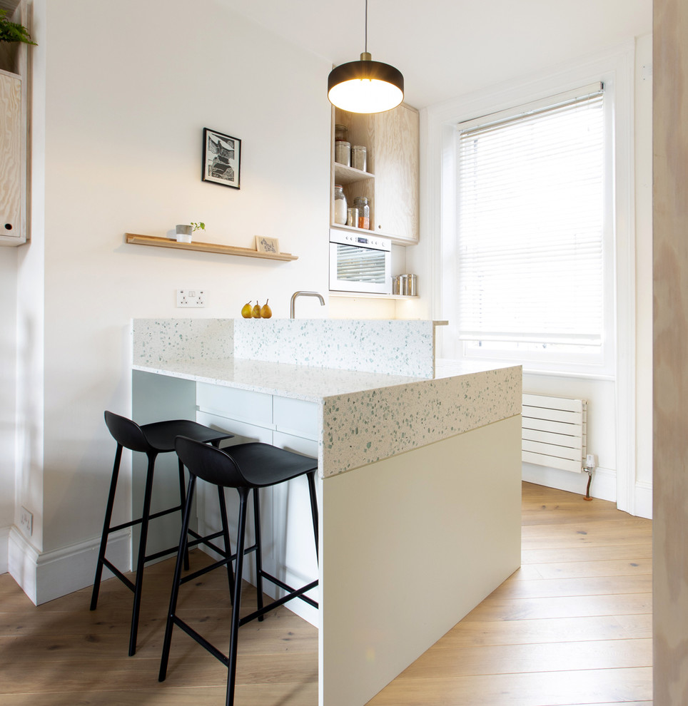 Small scandinavian open plan kitchen in London with a single-bowl sink, flat-panel cabinets, recycled glass benchtops, white splashback, stainless steel appliances, medium hardwood floors, with island and white benchtop.