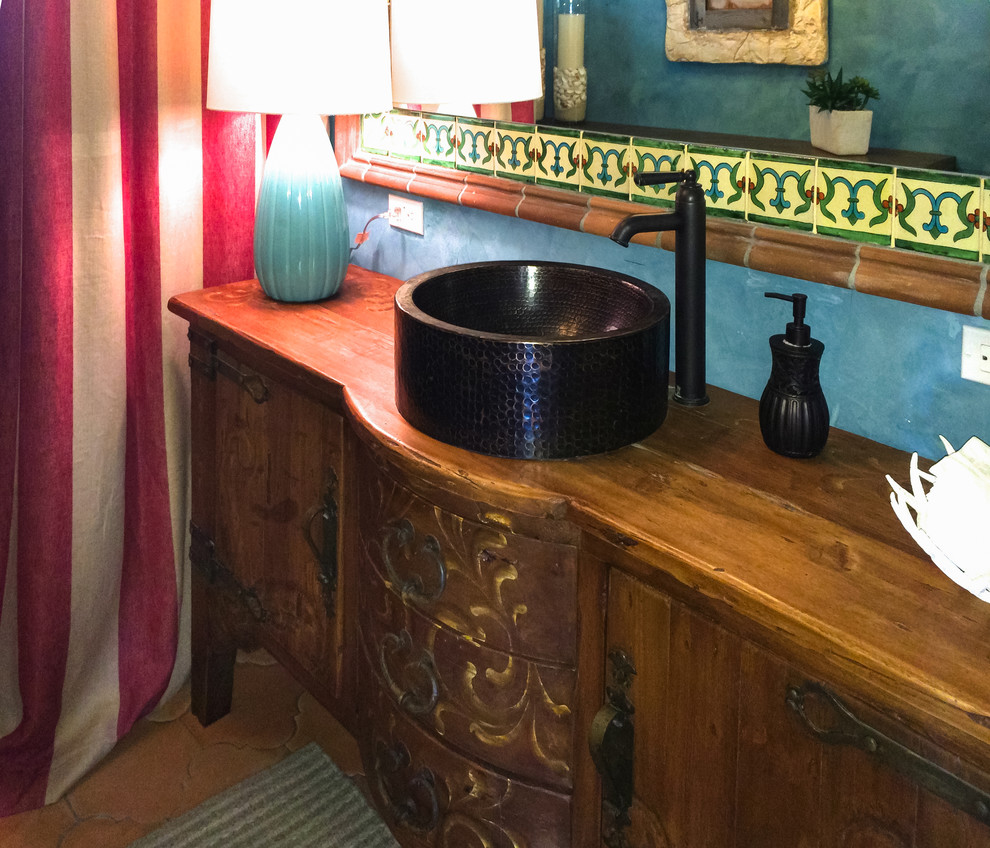 This is an example of a mid-sized mediterranean powder room in Other with furniture-like cabinets, medium wood cabinets, blue walls, terra-cotta floors, a vessel sink, wood benchtops and brown floor.