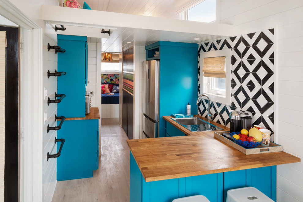 Small eclectic l-shaped open plan kitchen in Philadelphia with a drop-in sink, shaker cabinets, wood benchtops, ceramic splashback, stainless steel appliances, vinyl floors, a peninsula, timber, blue cabinets, multi-coloured splashback, beige floor and brown benchtop.
