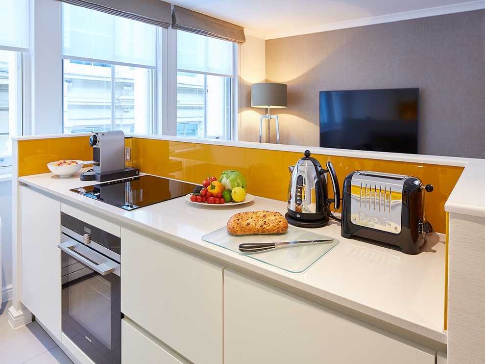 Contemporary open plan kitchen in London with flat-panel cabinets, white cabinets, orange splashback, glass sheet splashback and white benchtop.