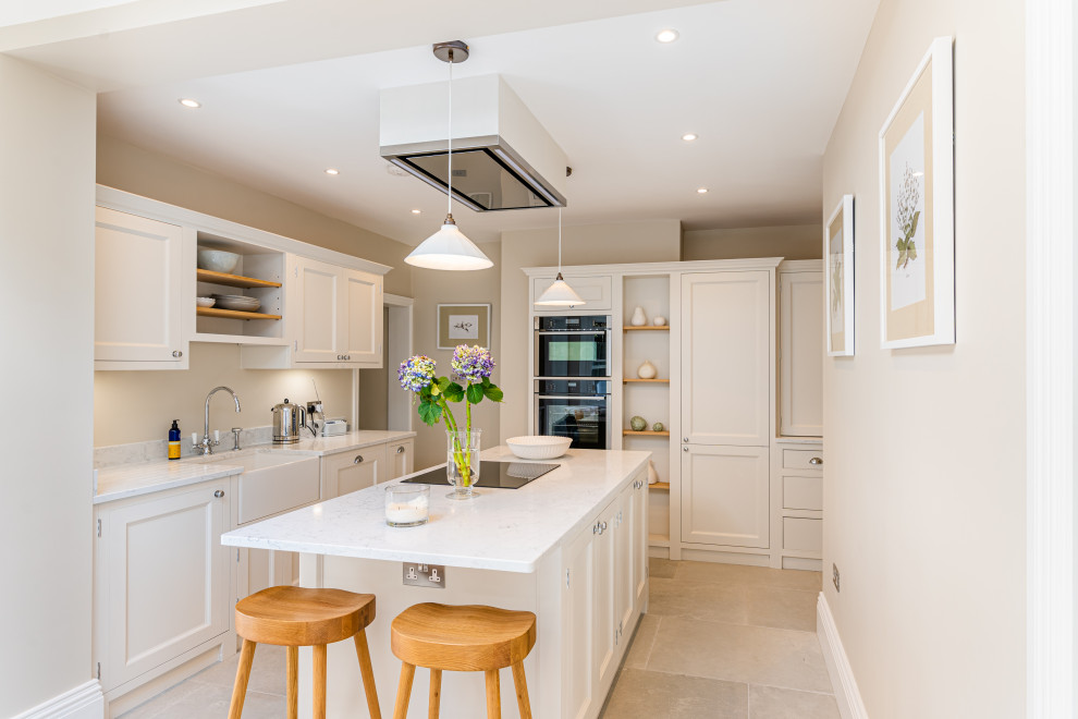 This is an example of a medium sized classic l-shaped open plan kitchen in London with a belfast sink, shaker cabinets, beige cabinets, composite countertops, black appliances, porcelain flooring, an island, grey floors and white worktops.