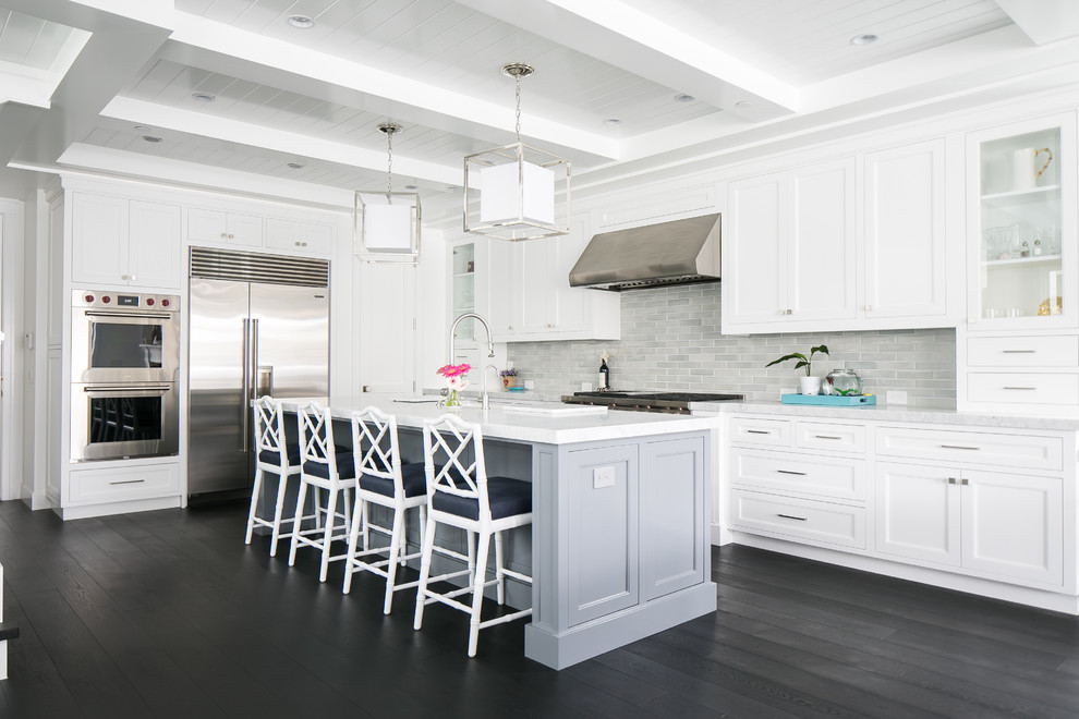 Photo of a large beach style l-shaped open plan kitchen in Orange County with white cabinets, marble benchtops, grey splashback, ceramic splashback, stainless steel appliances, dark hardwood floors, with island, shaker cabinets and a farmhouse sink.