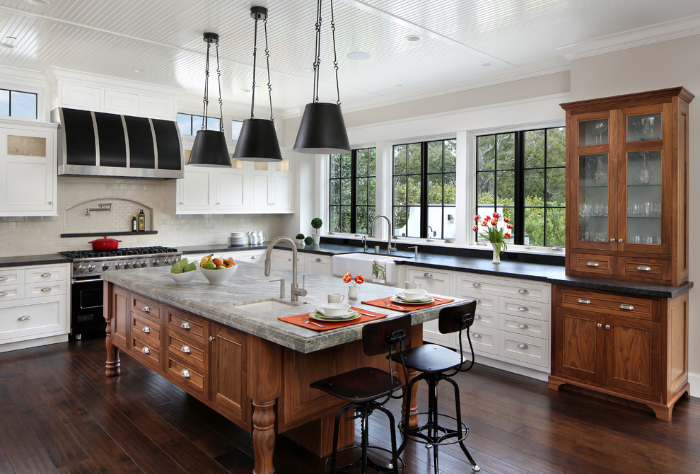 This is an example of a large traditional l-shaped open plan kitchen in San Francisco with a farmhouse sink, recessed-panel cabinets, white cabinets, white splashback, black appliances, dark hardwood floors, with island, granite benchtops, subway tile splashback and brown floor.