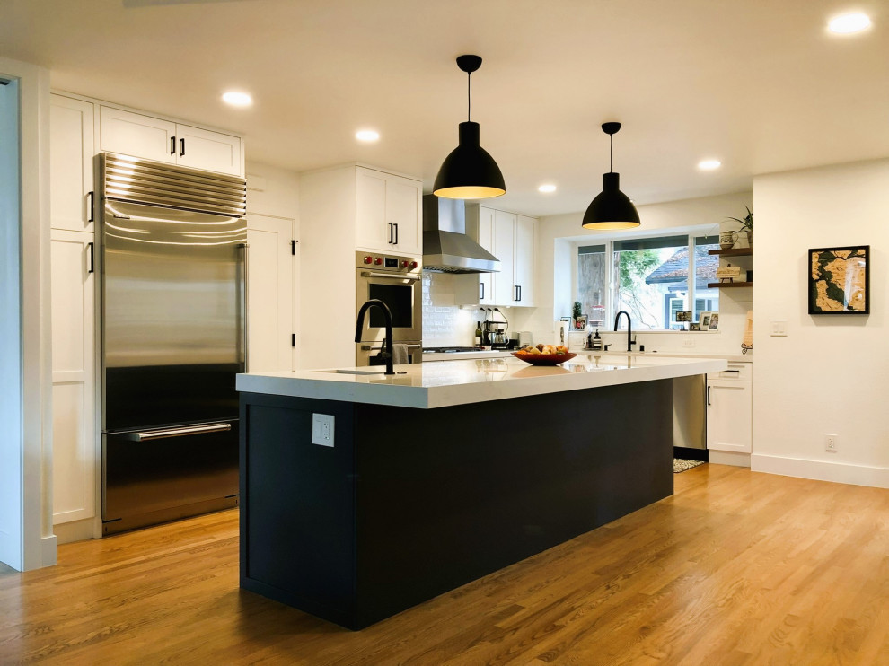 Photo of a mid-sized transitional l-shaped open plan kitchen in San Francisco with a single-bowl sink, shaker cabinets, black cabinets, quartz benchtops, white splashback, ceramic splashback, stainless steel appliances, medium hardwood floors, with island and white benchtop.