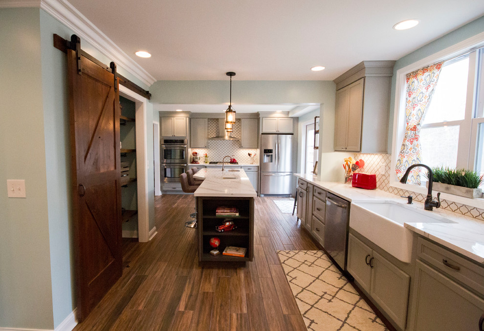 Large transitional l-shaped medium tone wood floor and brown floor enclosed kitchen photo in Cincinnati with a farmhouse sink, recessed-panel cabinets, gray cabinets, marble countertops, white backsplash, porcelain backsplash, stainless steel appliances and an island