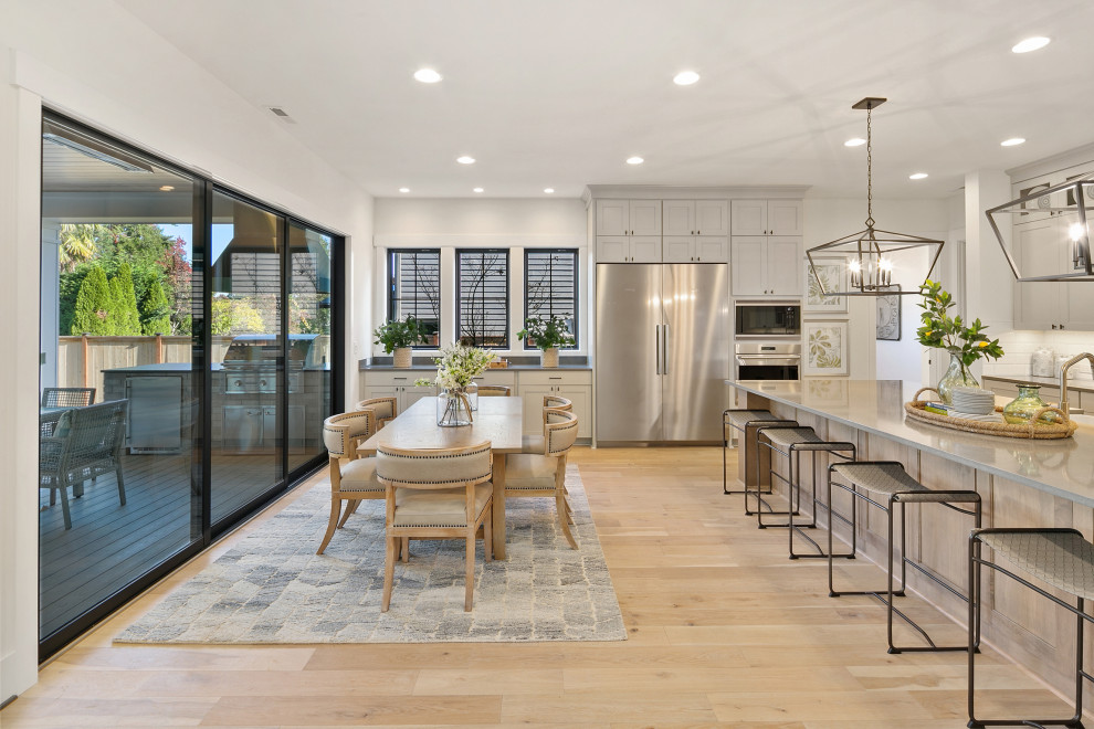 Photo of a large country l-shaped eat-in kitchen in Seattle with recessed-panel cabinets, grey cabinets, quartz benchtops, white splashback, stainless steel appliances, grey benchtop, a farmhouse sink, light hardwood floors, with island and beige floor.