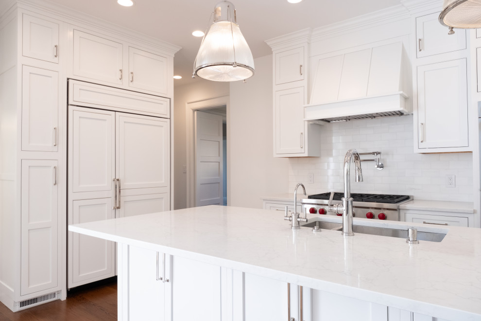 Photo of a mid-sized transitional l-shaped eat-in kitchen in New York with a double-bowl sink, beaded inset cabinets, white cabinets, quartz benchtops, white splashback, subway tile splashback, panelled appliances, medium hardwood floors, with island, brown floor and white benchtop.